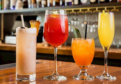 A row of colorful cocktails on a wooden bar, with bottles lined up behind the bar.