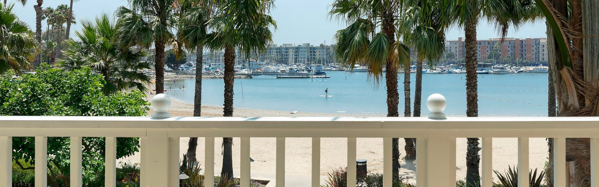 A beach view with palm trees, a white railing in the foreground, boats in the harbor, and a person paddleboarding on the water.