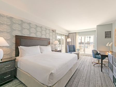 A hotel room with a king-sized bed, stylish decor, desk, and chairs. Large windows offer a view and natural light fills the space.