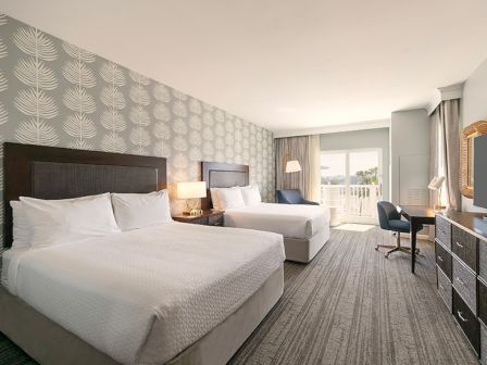 The image shows a hotel room with two beds, a patterned wall, a desk, a TV, and a window leading to a balcony.