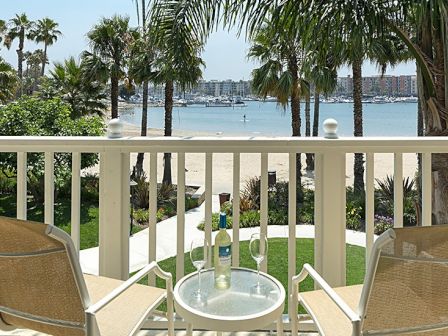 A balcony overlooking a beach with two chairs, a table, wine bottle, glasses, palm trees, and distant boats in the water.