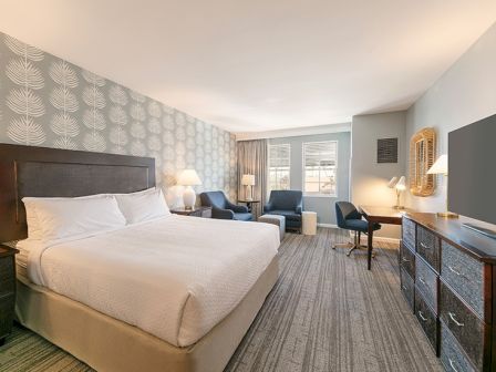 A hotel room with a bed, two chairs, a desk, and a TV, featuring patterned wallpaper and ample lighting.