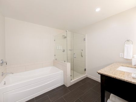 The image shows a modern bathroom with a bathtub, glass shower, and granite-topped vanity against white walls and dark flooring.