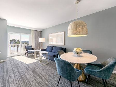 A modern living area features a blue sofa, round dining table with chairs, pendant light, and access to a balcony with natural light.