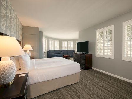 The image shows a modern hotel room with a bed, two lamps, a dresser, a TV, a sofa, and several windows with shutters.