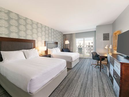 The image shows a hotel room with two beds, a desk, a TV, and a sliding door leading to a balcony, featuring modern decor.