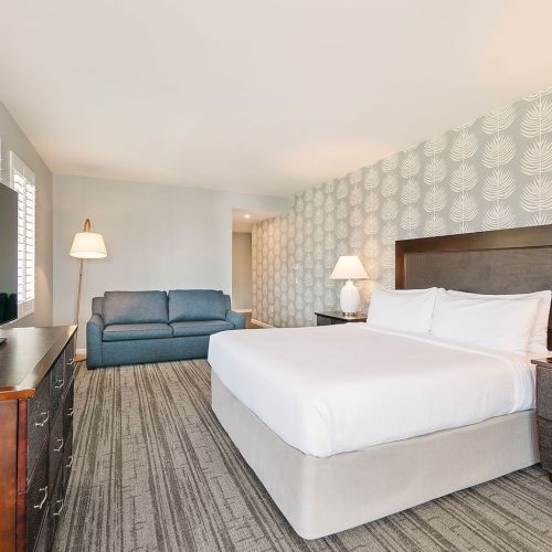 The image shows a hotel room with a bed, two nightstands with lamps, a TV, and a couch in a softly lit setting.