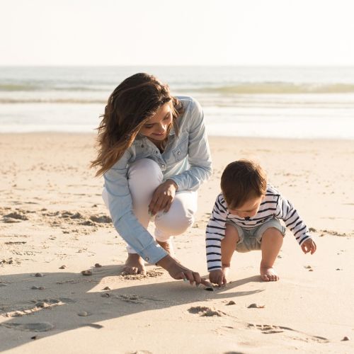 A woman and a child are playing on a sandy beach near the ocean, picking up objects and enjoying the sunny day.