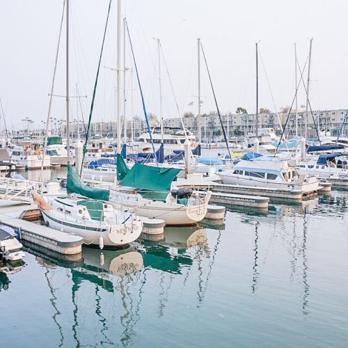 The image shows a marina with multiple sailboats docked, reflecting on the calm water. The sky looks clear, creating a serene scene.