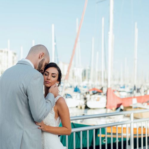 A couple embraces tenderly near a marina, with boats and masts in the background on a clear day.