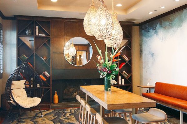 A stylish dining room with a wooden table, stools, pendant lights, abstract carpet, and wall art, with a mirror and bookshelf.