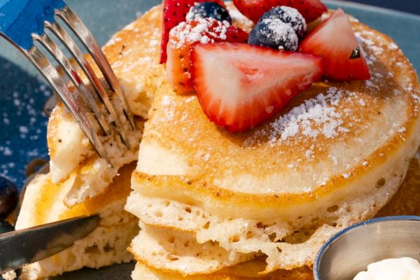 A stack of pancakes topped with strawberries and blueberries, accompanied by a small container of cream on a plate.