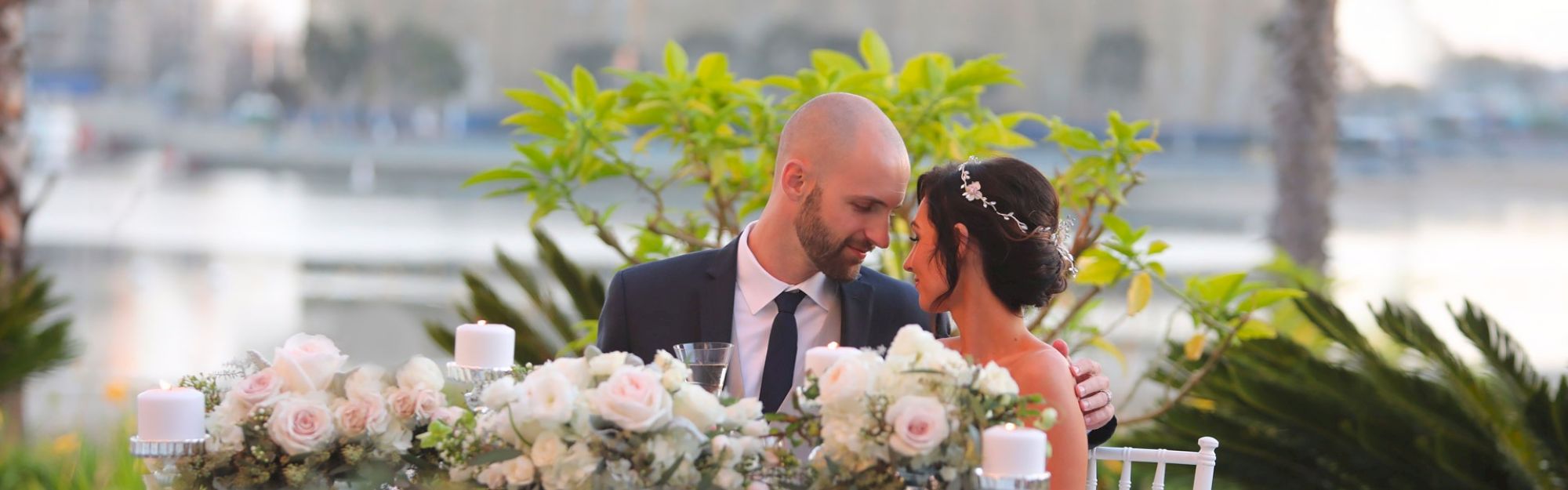 A couple sits close at a floral wedding table, gazing at each other with a lakeside backdrop and soft pink roses lining the centerpiece.