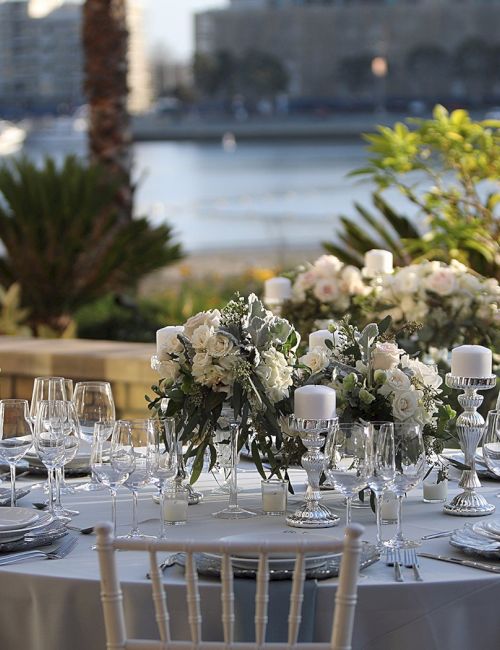 A formal outdoor table setting by the water with white flowers, glassware, and candles, ready for a wedding or elegant event.
