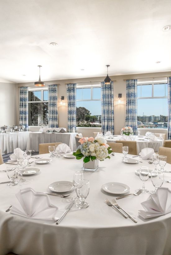 A bright, elegant dining room with round tables dressed in white linens, place settings, and floral centerpieces, ready for a formal meal.