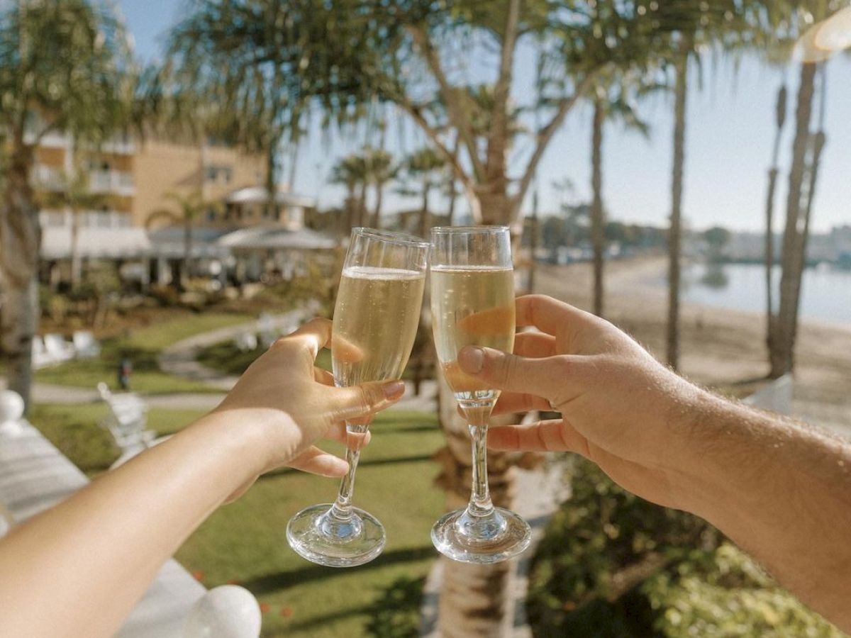 A sunny resort scene with people clinking champagne glasses on a balcony, palm trees, and a waterfront view.