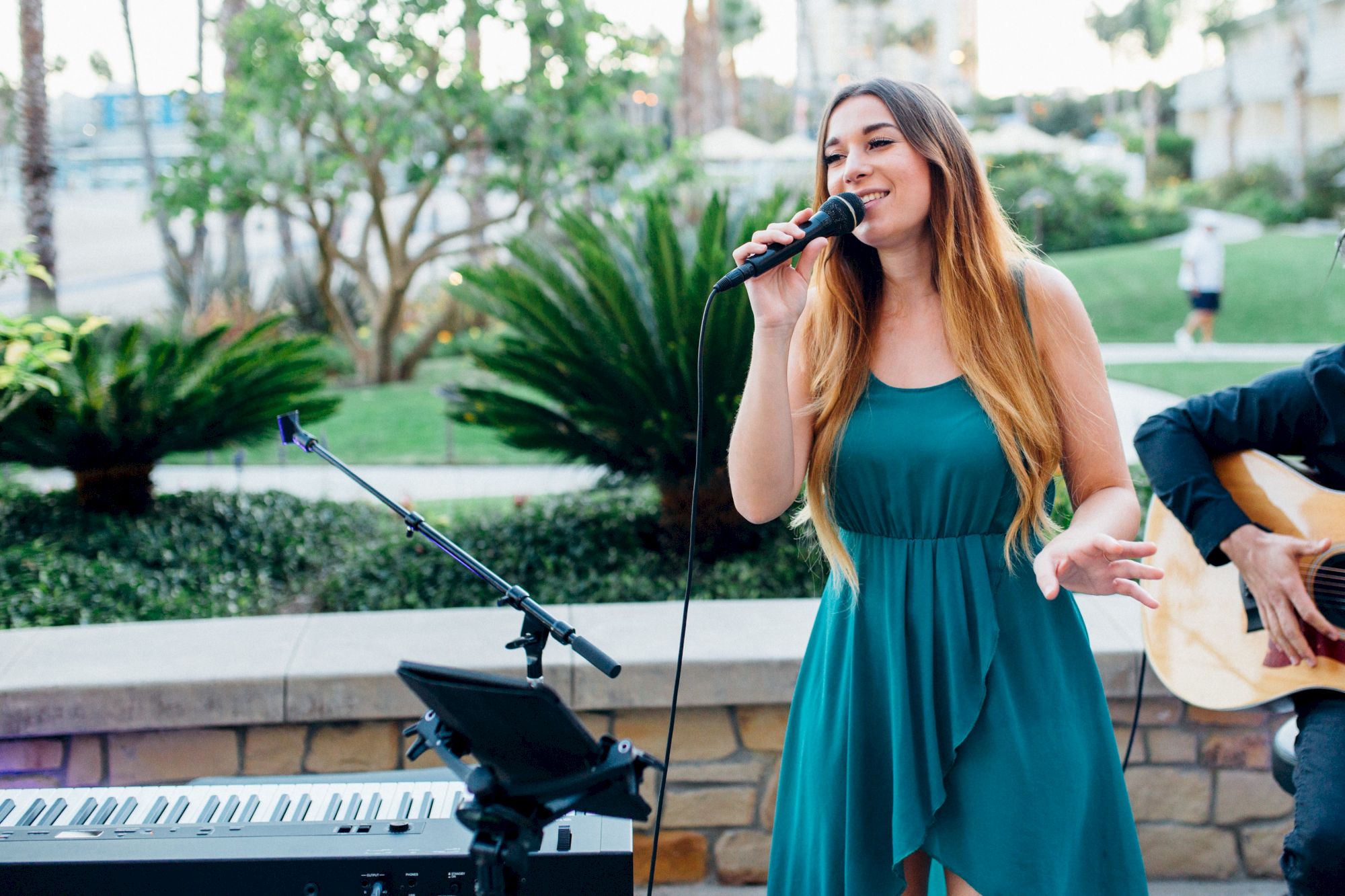 A female singer in a teal dress performs outdoors with a mic, a guitarist beside her, and a keyboard setup nearby, in a sunny garden setting.