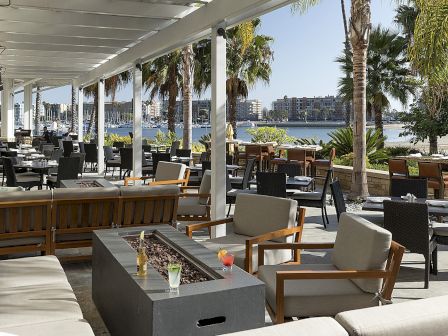A stylish outdoor cafe by the water with lounge seating, wooden chairs, palm trees, and a shaded patio overlooking a lake or bay.