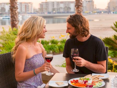 A couple dining outdoors by the beach, sharing wine and plates of snacks under a warm sunset glow.