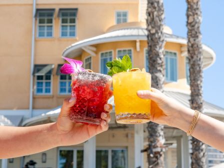 Two colorful drinks clink in front of a sunny hotel backdrop; one red with a flower, the other orange; cheers to sunny vibes.