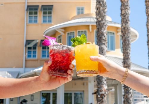 Two hands clink colorful cocktails in front of a sunny hotel, with garnishes and a bright, tropical vibe. End with a period.