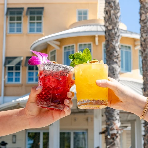 Two hands clink colorful cocktails in front of a sunny hotel, with garnishes and a bright, tropical vibe. End with a period.