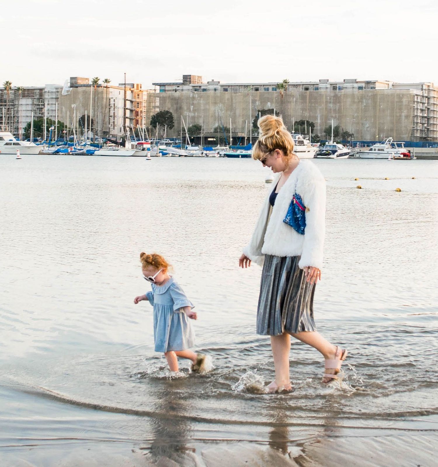 Two people walking along the shallow harbor water, a child in a blue outfit and an adult guiding them, with boats and buildings in the background.