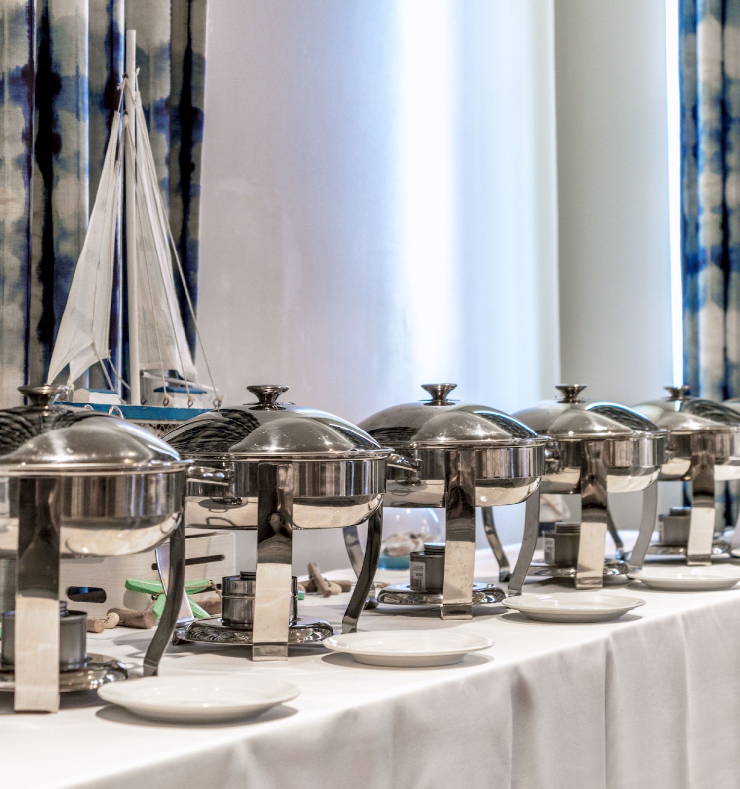 A row of shiny chafing dishes on a buffet table with a model sailboat centerpiece in front of striped curtains, ready for serving.